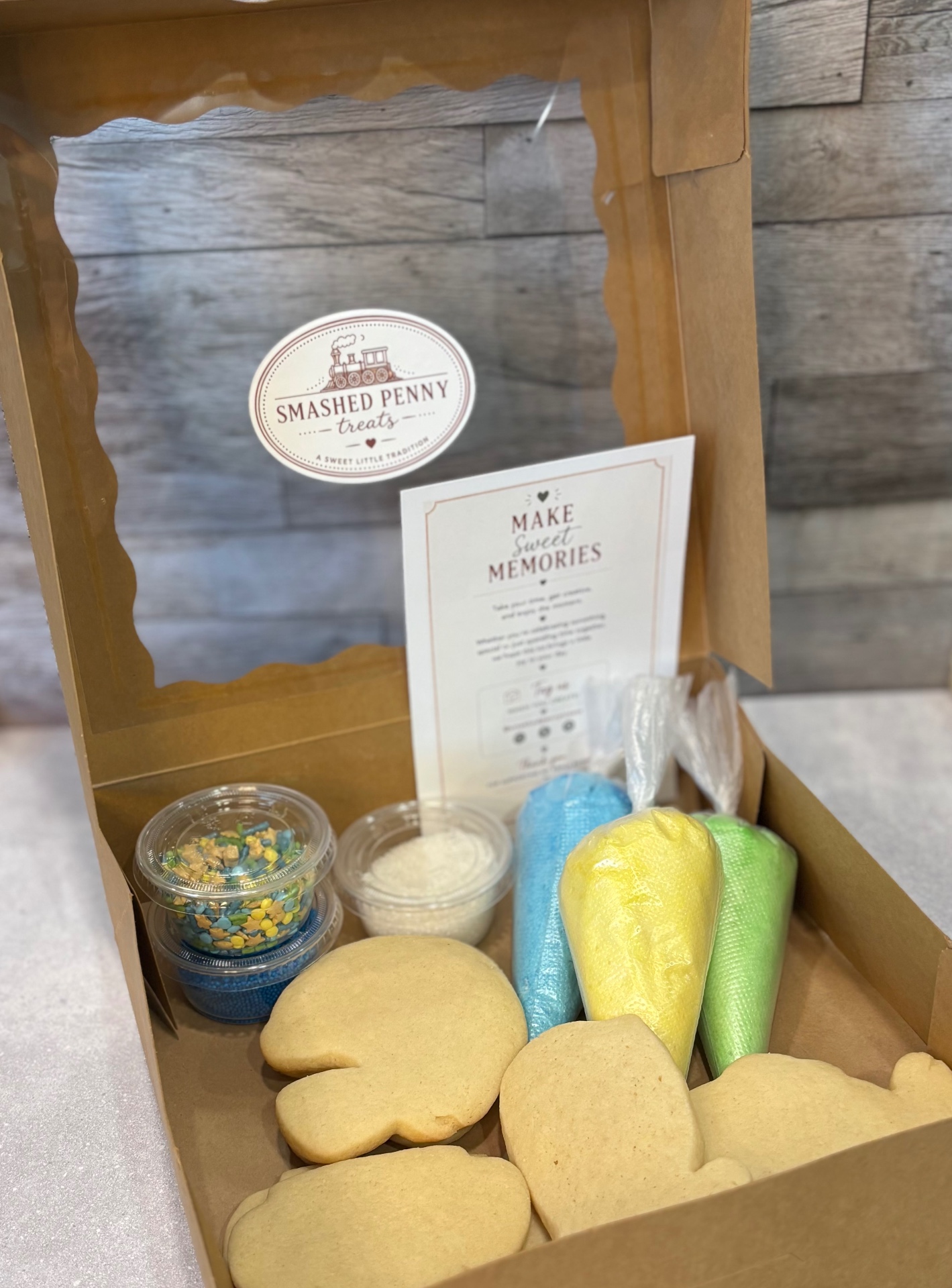 An open kraft cookie kit box with the Smashed Penny Treats logo, fresh sugar cookies, frosting bags, and sprinkles inside